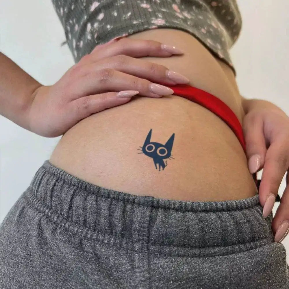 Small dark blue coal ball cat tattoo with luminous eyes on hip of person in gray sweatpants